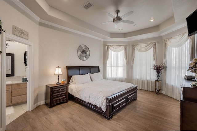 bedroom with a tray ceiling, crown molding, visible vents, ensuite bathroom, and light wood-type flooring