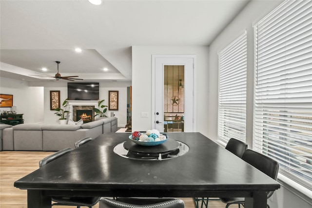 dining space with a warm lit fireplace, a raised ceiling, a ceiling fan, light wood-style floors, and recessed lighting