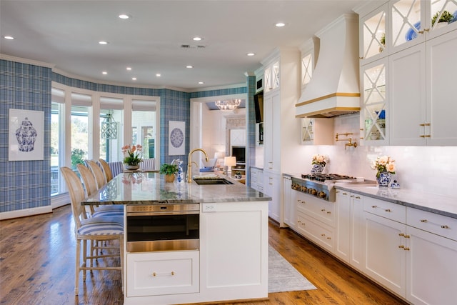 kitchen with a center island with sink, appliances with stainless steel finishes, glass insert cabinets, premium range hood, and a sink