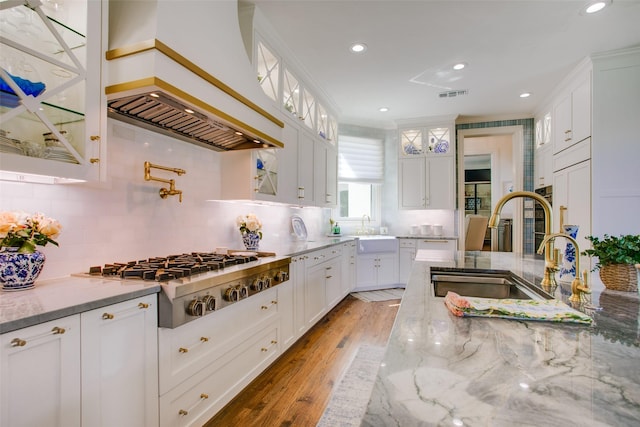 kitchen featuring stainless steel gas cooktop, glass insert cabinets, white cabinetry, light stone countertops, and premium range hood