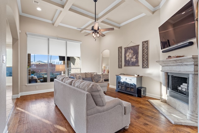 living area featuring arched walkways, a fireplace with raised hearth, coffered ceiling, baseboards, and dark wood-style floors