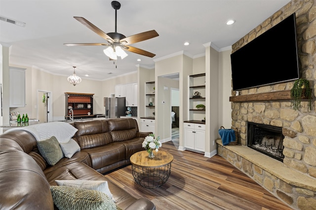 living area with visible vents, crown molding, a stone fireplace, ceiling fan with notable chandelier, and wood finished floors