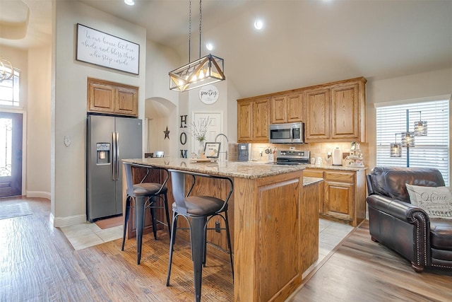 kitchen with arched walkways, light stone counters, a kitchen island with sink, stainless steel appliances, and decorative backsplash