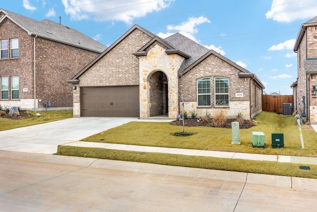 french country inspired facade with a shingled roof, concrete driveway, an attached garage, cooling unit, and a front lawn