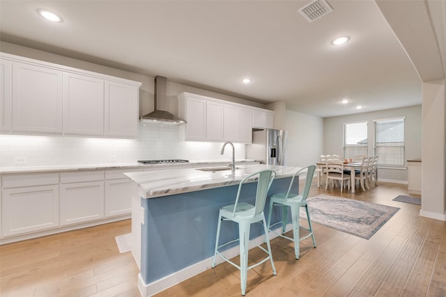 kitchen featuring white cabinets, an island with sink, wall chimney exhaust hood, stainless steel appliances, and light wood-type flooring