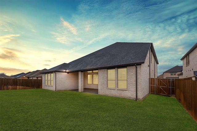 back of house at dusk featuring a yard, a fenced backyard, roof with shingles, and brick siding