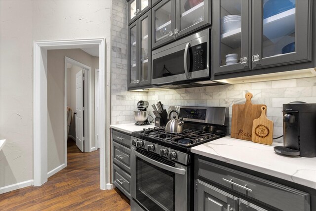 kitchen featuring light stone counters, dark wood-style flooring, decorative backsplash, appliances with stainless steel finishes, and glass insert cabinets