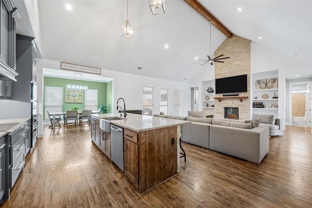 kitchen featuring light stone counters, hanging light fixtures, stainless steel dishwasher, a kitchen island with sink, and a sink