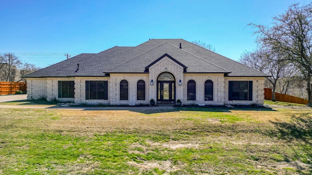 french country home with a shingled roof, brick siding, fence, and a front lawn