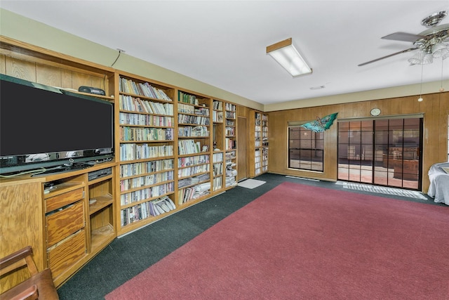 living area featuring ceiling fan, dark colored carpet, bookshelves, and wooden walls