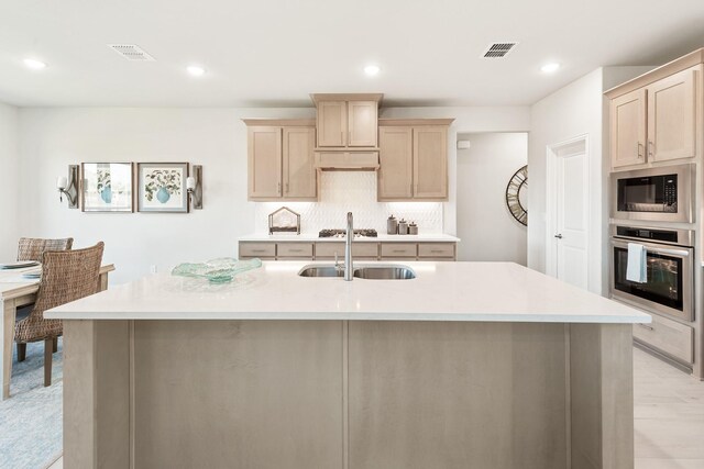 kitchen featuring a center island with sink, appliances with stainless steel finishes, light countertops, light brown cabinetry, and a sink