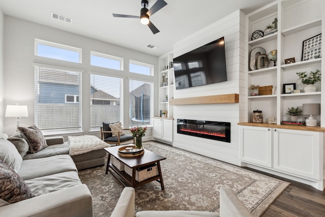 living room featuring built in features, a glass covered fireplace, dark wood-style flooring, and visible vents