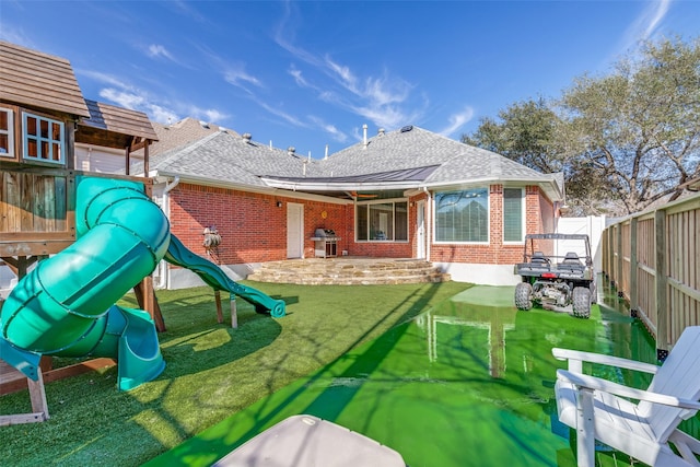 rear view of property with a shingled roof, fence, a yard, a playground, and brick siding