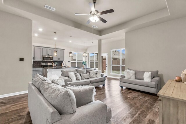 living area with ceiling fan, dark wood finished floors, visible vents, baseboards, and a raised ceiling