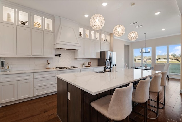 kitchen featuring light stone counters, glass insert cabinets, custom exhaust hood, a center island with sink, and pendant lighting