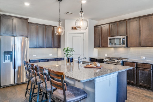 kitchen featuring dark wood finished floors, appliances with stainless steel finishes, light stone countertops, a kitchen island with sink, and a sink