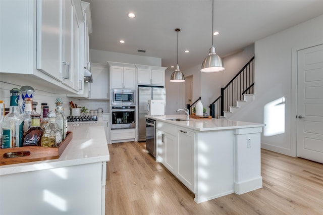 kitchen featuring an island with sink, stainless steel appliances, light wood-type flooring, white cabinetry, and pendant lighting