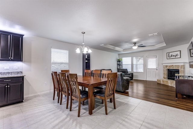 dining space featuring light tile patterned floors, a stone fireplace, ceiling fan with notable chandelier, visible vents, and a tray ceiling