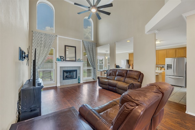 living room featuring a tile fireplace, a towering ceiling, a ceiling fan, dark wood finished floors, and crown molding