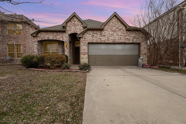 french provincial home with a garage, a shingled roof, brick siding, driveway, and stone siding