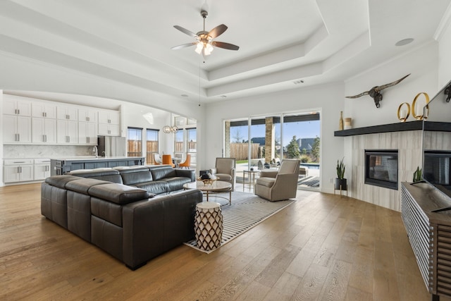 living room with ceiling fan with notable chandelier, visible vents, light wood-style floors, a raised ceiling, and a glass covered fireplace