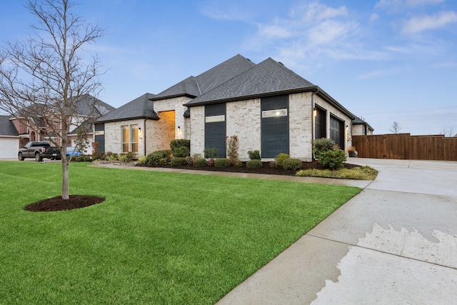 french country style house with concrete driveway, roof with shingles, fence, a front lawn, and brick siding