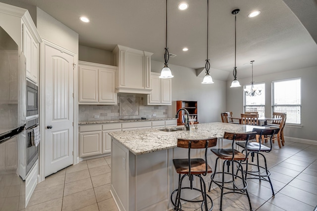 kitchen featuring decorative light fixtures, a center island with sink, white cabinets, a sink, and light stone countertops