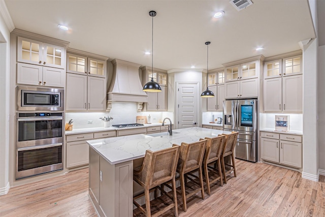 kitchen featuring appliances with stainless steel finishes, premium range hood, a center island with sink, and glass insert cabinets