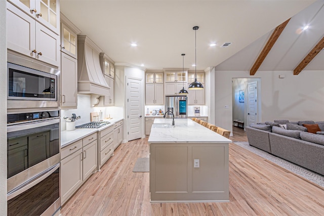 kitchen with stainless steel appliances, open floor plan, custom exhaust hood, an island with sink, and glass insert cabinets