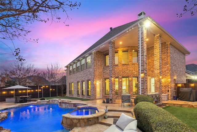 back of house at dusk featuring a patio, brick siding, fence, and a pool with connected hot tub