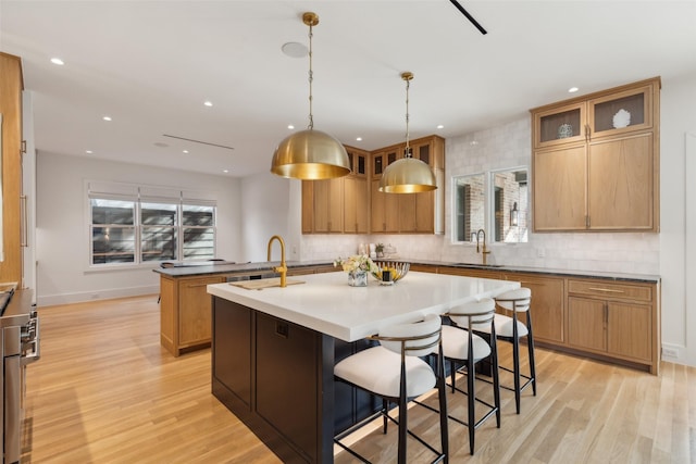 kitchen with an island with sink, glass insert cabinets, light wood-style floors, pendant lighting, and a sink