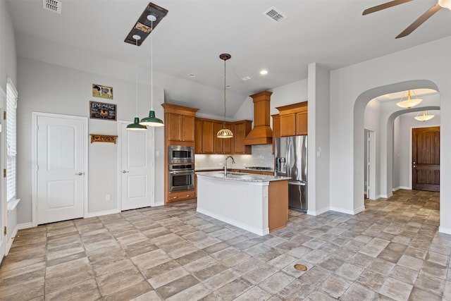 kitchen with pendant lighting, custom range hood, appliances with stainless steel finishes, a kitchen island with sink, and light stone countertops