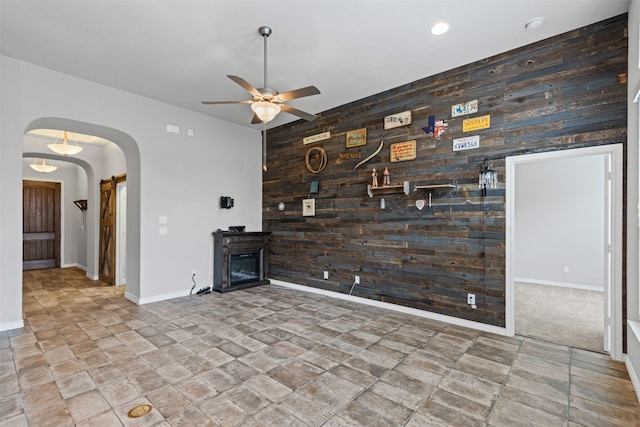 unfurnished living room featuring wooden walls, stone finish floor, arched walkways, an accent wall, and ceiling fan