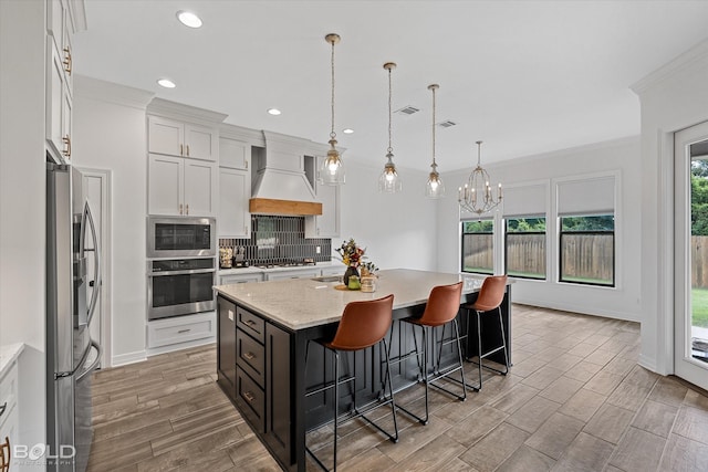 kitchen featuring pendant lighting, custom exhaust hood, appliances with stainless steel finishes, a kitchen island with sink, and white cabinetry