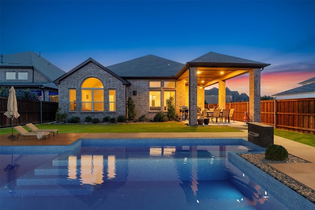 back of house at dusk featuring a fenced in pool, brick siding, roof with shingles, a patio area, and a fenced backyard