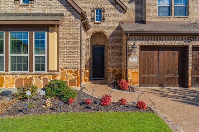 view of exterior entry with stone siding, concrete driveway, brick siding, and roof with shingles