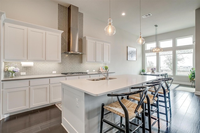 kitchen with light countertops, white cabinetry, a sink, wall chimney range hood, and an island with sink