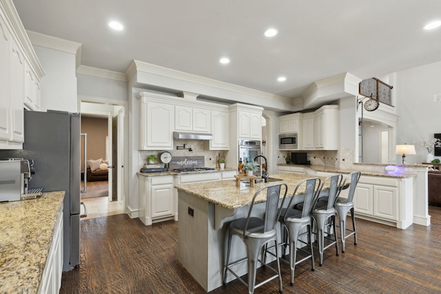 kitchen with backsplash, under cabinet range hood, a center island with sink, appliances with stainless steel finishes, and a kitchen breakfast bar