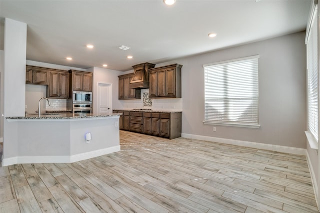 kitchen featuring light stone counters, stainless steel appliances, light wood-type flooring, tasteful backsplash, and custom range hood