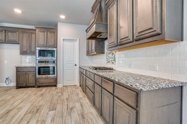 kitchen featuring stainless steel appliances, light wood finished floors, stone countertops, and custom range hood