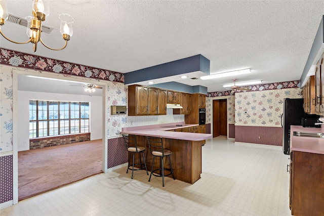 kitchen with wainscoting, a textured ceiling, a peninsula, a kitchen bar, and wallpapered walls