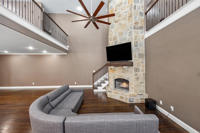 living room featuring dark wood-style floors, a towering ceiling, ceiling fan, a stone fireplace, and baseboards