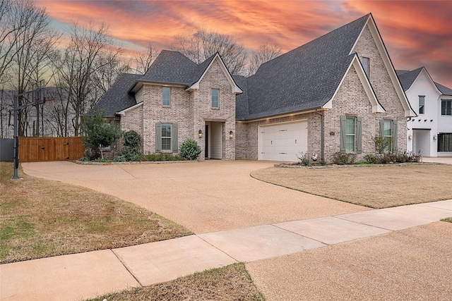 french country style house with driveway, a garage, a shingled roof, a gate, and brick siding