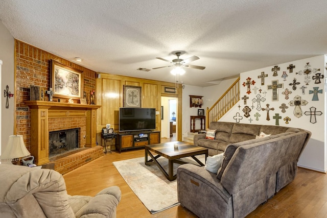 living room with a textured ceiling, light wood-style flooring, visible vents, stairs, and a brick fireplace