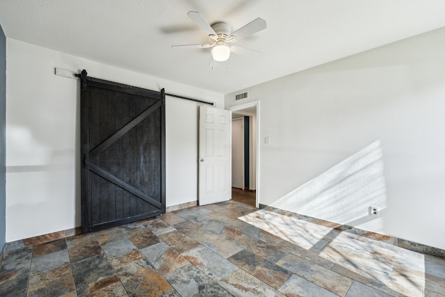 unfurnished bedroom featuring a barn door, visible vents, baseboards, ceiling fan, and stone finish floor