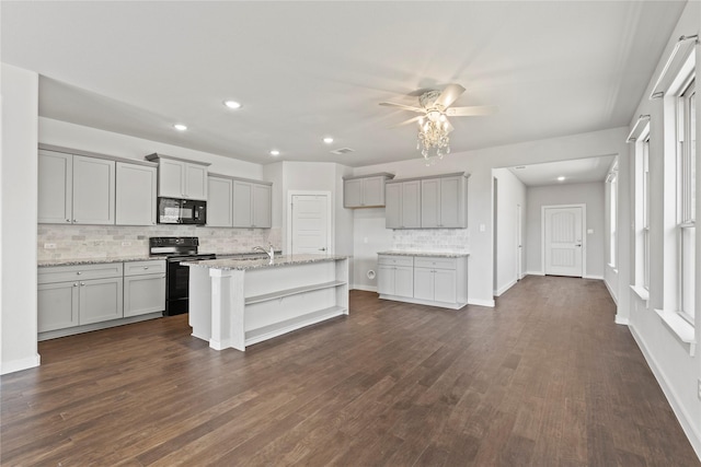 kitchen featuring light stone counters, dark wood-style flooring, a center island with sink, gray cabinets, and black appliances