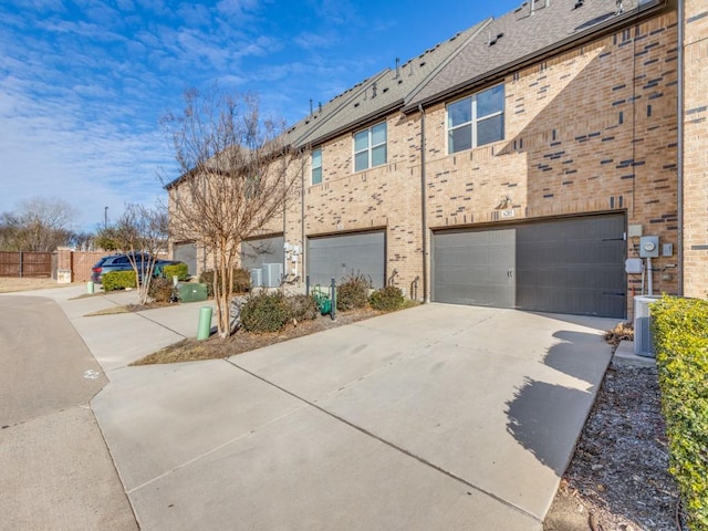 exterior space with central AC unit, an attached garage, brick siding, driveway, and roof with shingles