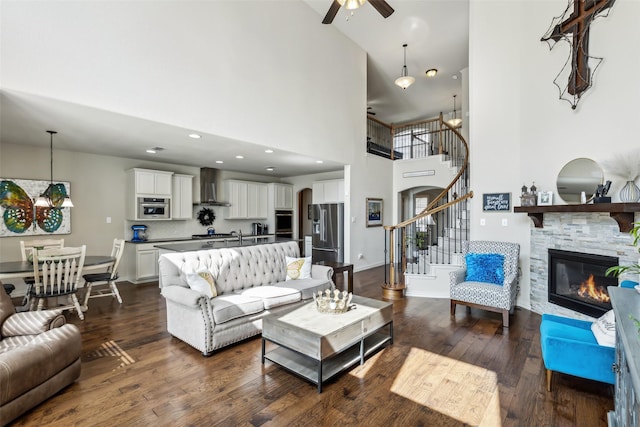 living room featuring a stone fireplace, ceiling fan with notable chandelier, baseboards, stairs, and dark wood-style floors