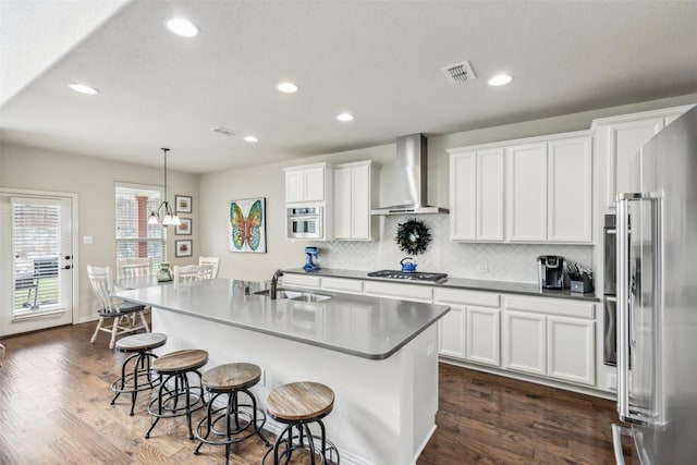 kitchen featuring white cabinets, an island with sink, decorative light fixtures, stainless steel appliances, and wall chimney range hood