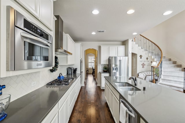 kitchen featuring arched walkways, stainless steel appliances, a sink, white cabinetry, and wall chimney exhaust hood